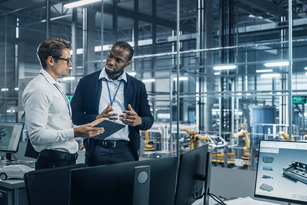 Two men stand in a manufacturing testing facility. They are looking at each other and appear to be trying to solve a problem. On the left is a Black male with black hair and a short beard, wearing a light blue, open-collared shirt, a navy blue jacket, dark blue pants with a black belt, and a blue lanyard around his neck. He holds a pencil in his left hand, and both hands are raised slightly and open in toward himself On the right stands a white man with dark brown hair and glasses.